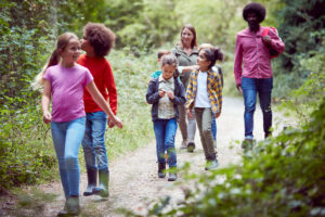 Adult Team Leaders With Group Of Children At Outdoor Activity Camp Walking Through Woodland